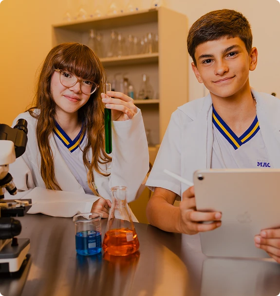 Dois alunos do Colégio Magnum em atividade de laboratório: ela observa um tubo de ensaio, ele anota num tablet, com microscópio e vidraria na mesa