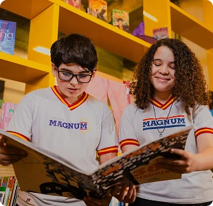 Dois alunos do Colégio Magnum lendo um livro grande juntos, em frente às estantes coloridas
