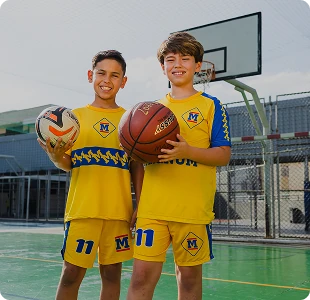 Dois alunos do Colégio Magnum de uniforme esportivo amarelo, com bolas de futebol e basquete na quadra
