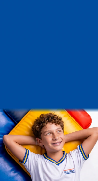 Aluno do Ensino Fundamental do Colégio Magnum deitado num colchonete colorido, com as mãos atrás da cabeça, usando o uniforme da escola